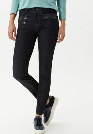 Jean en denim foncé avec une coupe slim, dotés de poches zippées décoratives et de coutures contrastantes ; associés à des baskets navy.