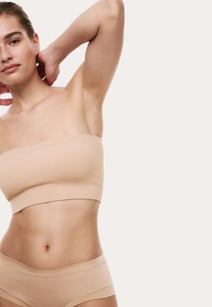 Kvinde iført beige stropløs bandeau-top og matchende undertøj, positur med armene løftet mod en ensartet lys baggrund.