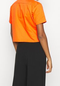 Orange cropped short-sleeve shirt with a collar, featuring a smooth texture. Paired with black wide-leg pants.
