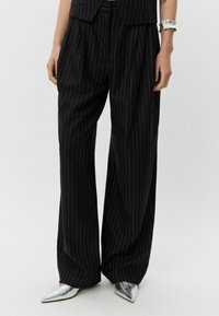 Black pinstriped trousers with a loose fit, featuring pleats and a high waist, paired with silver pointed-toe shoes.