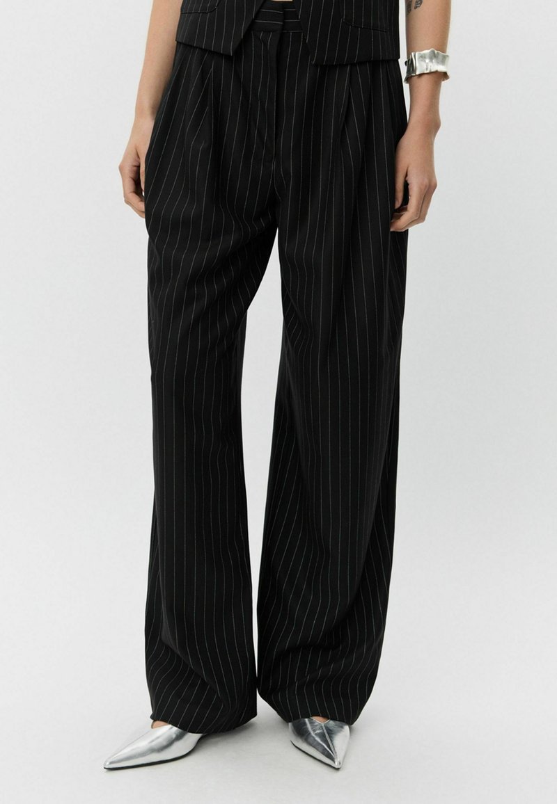 Black pinstriped trousers with a loose fit, featuring pleats and a high waist, paired with silver pointed-toe shoes.