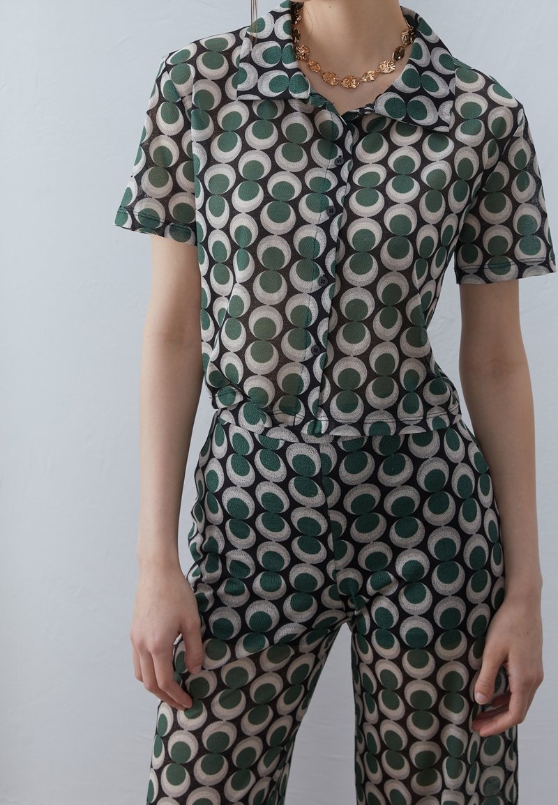 Personne portant une chemise à manches courtes boutonnée et un pantalon assortis à motifs verts et beiges, avec un collier chaîne en or.