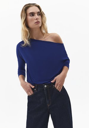 Kobieta o blond falowanych włosach, ubrana w niebieską bluzkę z odkrytymi ramionami i ciemne dżinsy, z rękami w kieszeniach, patrząca w bok.