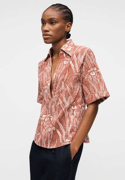 Femme aux cheveux tressés portant un chemisier à manches courtes de couleur rouille avec un motif floral abstrait blanc et un pantalon sombre, debout avec une main dans la poche.