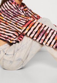 Henkilö, joka käyttää moniväristä raidallista neulepaitaa, jossa on vaaleanpunaisia, oransseja ja mustia raitoja, yhdistettynä vaaleisiin farkkuihin, istuu polvet ylhäällä.