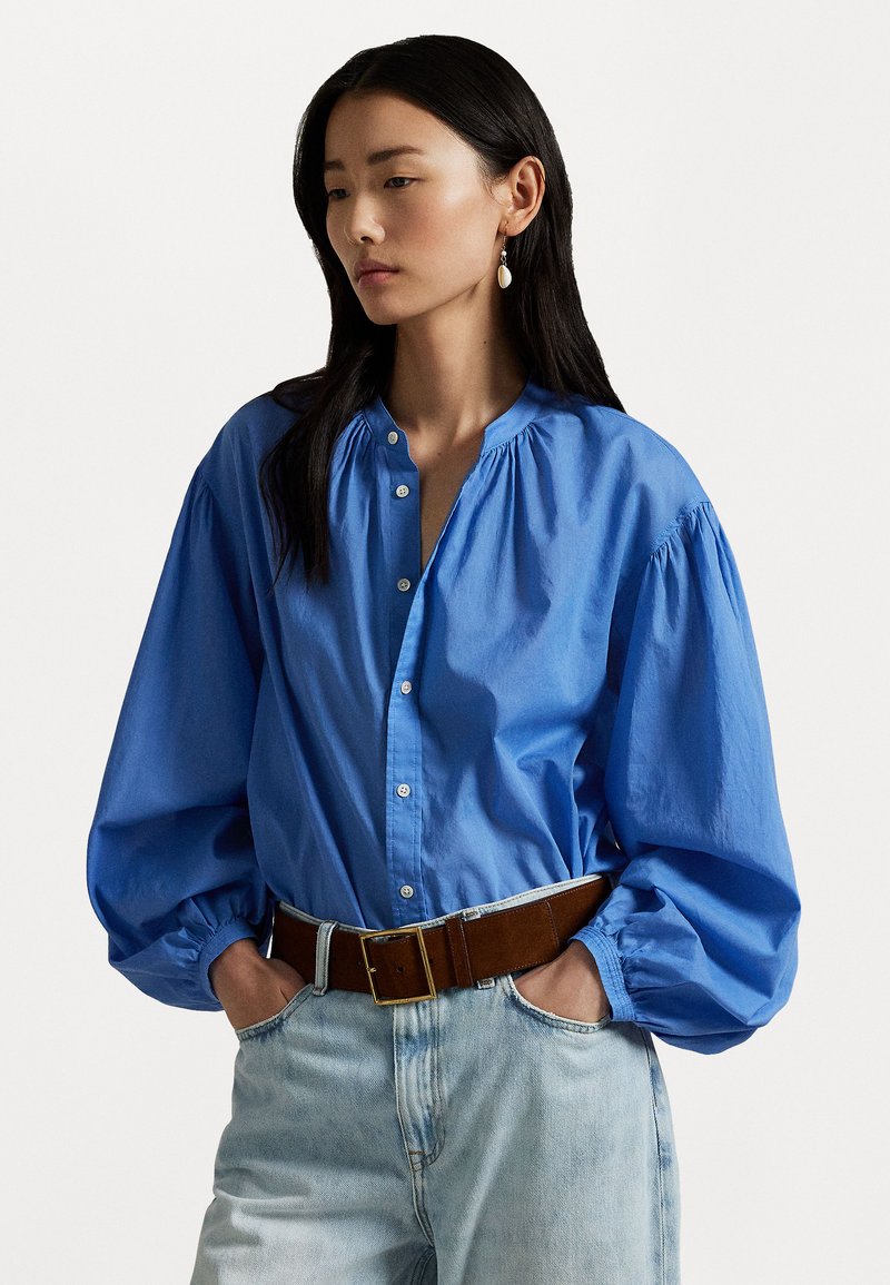 Blue cotton blouse with puffed sleeves and a button-down front, paired with high-waisted light denim and a brown belt.