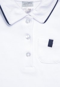 Hvit polo skjorte for babyer, med marineblå krage, tre klare knapper og en liten brystlomme med marineblå detalj.