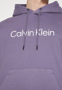 Felpa con cappuccio viola con logo bianco "Calvin Klein", cordini regolabili e tasca a marsupio frontale. Tessuto morbido.
