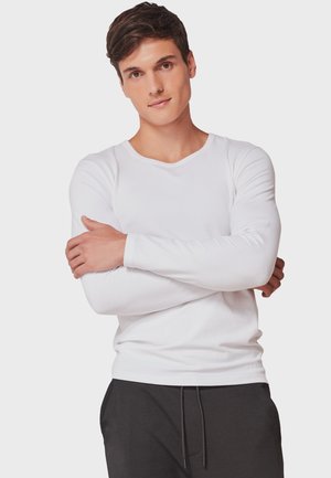 Camiseta blanca de manga larga con cuello redondo, fabricada en una tela suave. El modelo posa con los brazos cruzados, vistiendo pantalones gris oscuro.