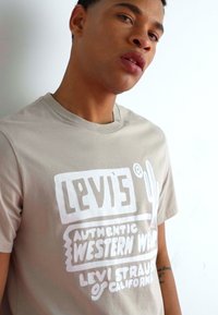 Camiseta de manga corta color beige con texto gráfico en blanco que incluye "Levi's," "Vestimenta Occidental Auténtica," y un diseño de cactus en la parte frontal.