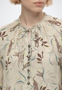 Gros plan d'un chemisier beige clair avec des motifs tigre et feuilles marron, un col orné de dentelle, une fermeture à nouer et des boutons métalliques décoratifs.