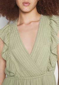 Vestido verde claro con una tela texturizada, que presenta mangas con volantes y un escote en V profundo, cintura elástica y silueta fluida.