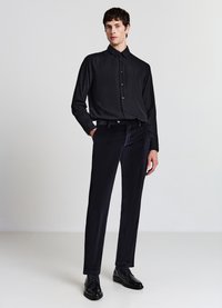 Camicia nera a bottoni con righe verticali, abbinata a pantaloni di velluto scuro e scarpe nere lucide, in piedi contro uno sfondo neutro.