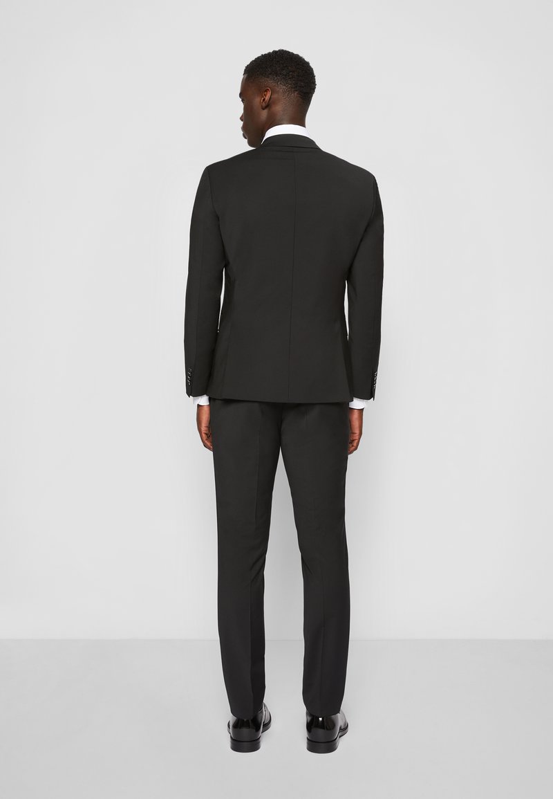 Zwart pak met een getailleerde pasvorm, voorzien van een enkele split, recht gesneden broek en glanzende zwarte schoenen. Witte overhemd bij de kraag.