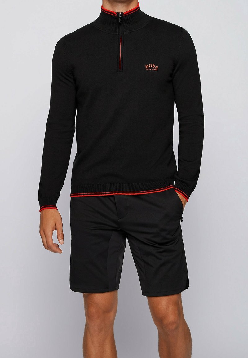 Homme portant un pull noir à manches longues avec fermeture éclair au quart, bordure rouge et logo BOSS, associé à un short noir à hauteur de genou, debout devant un fond uni.