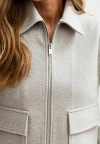 Veste en mélange de laine grise avec fermeture éclair, col pointu et deux grandes poches à rabat. Texture douce et coupe ajustée.