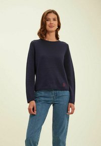 Pull en maille bleu marine à manches longues et col rond, avec un logo rouge brodé à l'ourlet, assorti à un jean bleu clair.