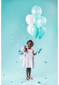 Ein Kind in einem weißen Kleid hält aqua- und weiße Ballons. Sie hat blaue Federakzente und ist von buntem Federkonfetti umgeben.