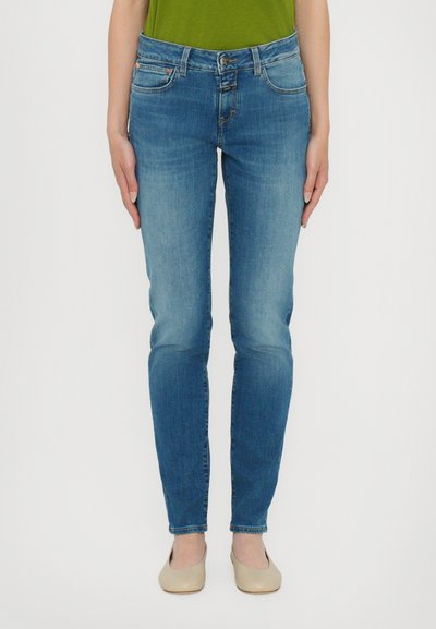 Jean en denim bleu skinny avec une taille mi-haute, design à cinq poches, délavage clair et détails de couture subtils. Portés avec des chaussures beiges.