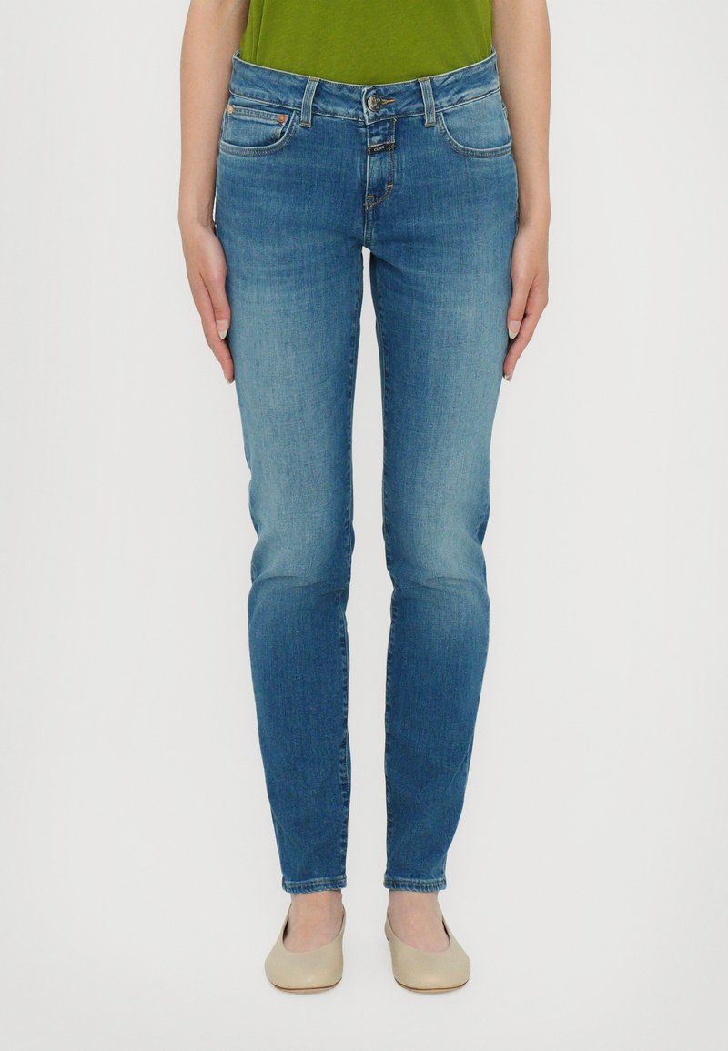 Tynne blå denimjeans med midjehøyde, fem lommer, lys vask og subtile sømdetaljer. Brukt med beige sko.