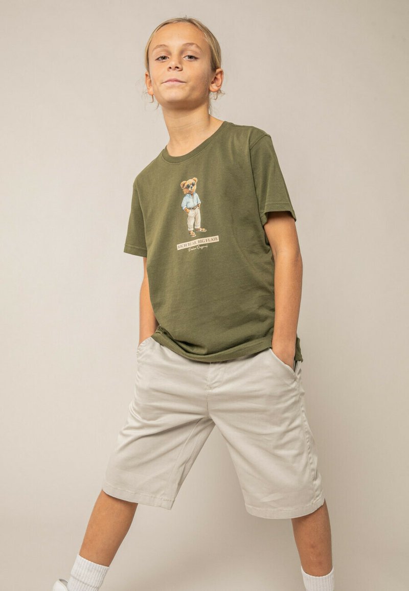 Enfant debout, les mains dans les poches, portant un t-shirt vert olive avec un motif d'ours et un short beige, sur fond beige uni.