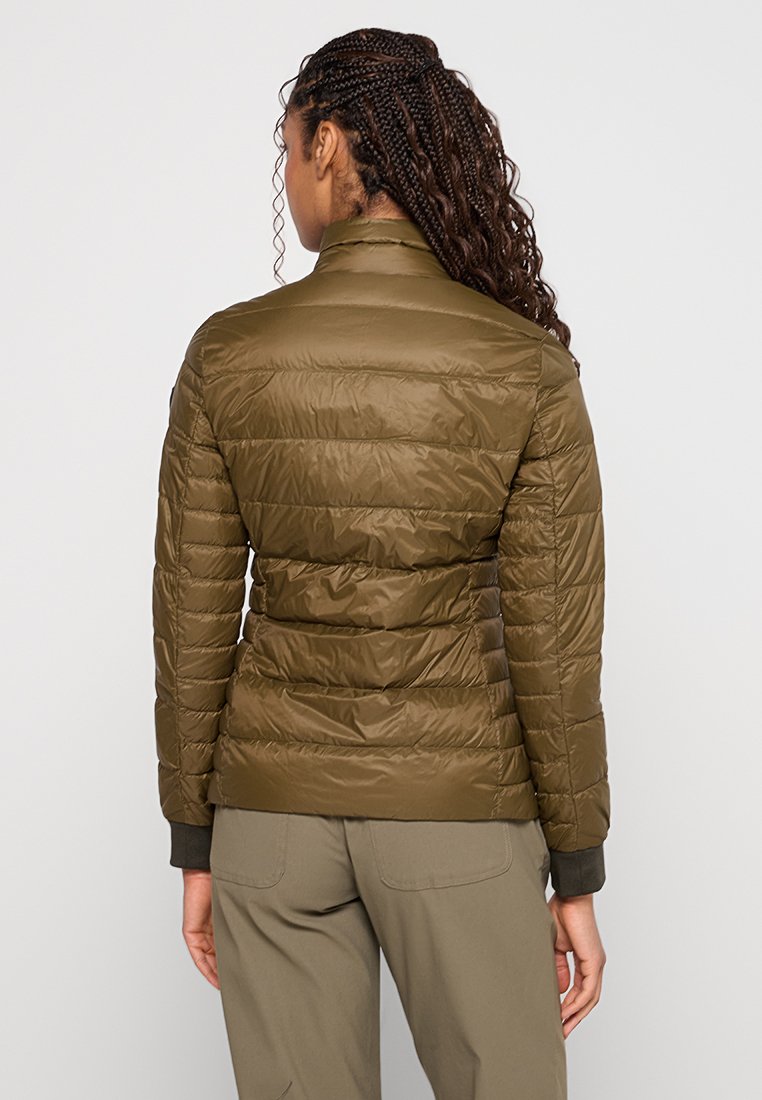 Frau mit geflochtenem Haar, die eine olivgrüne Steppjacke und passende Hose trägt, steht mit dem Rücken zu einem schlichten Hintergrund.