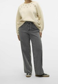 Sweatshirt beige chiaro con dettagli in borchie, abbinato a jeans grigi a vita alta e gamba larga e sneakers grigie con lacci neri.