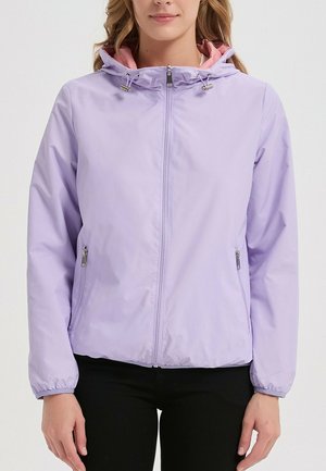 Femme portant une veste à capuche violet clair avec une fermeture éclair à l'avant et des poches latérales zippées, associée à un pantalon noir.