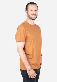 Bruine katoenen t-shirt met een ronde hals, korte mouwen en een klein logo op de linkerborst. Gecombineerd met donkere jeans, casual gedragen.