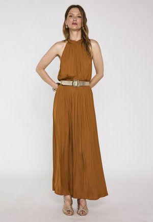 Femme debout dans une robe longue marron sans manches, ceinture dorée et sandales dorées à talons hauts, faisant face à l'avant sur un fond uni.