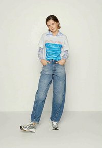 Pessoa de pé com as mãos nos bolsos, vestindo uma camisa de manga longa com padrão azul por baixo de uma camiseta gráfica branca e uns jeans azuis largos com ténis.