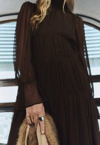 Vestido maxi plisado marrón con mangas largas transparentes y cuello alto. La modelo sostiene un bolso de mano beige acentuado con piel, que muestra anillos de plata.