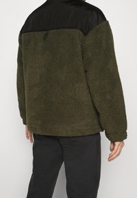 Olijf groene fleecejas met een zwart satijnen schouderpaneel, relaxte pasvorm en een gestructureerd oppervlak. Kenmerkt polsboorden en een lusdetail aan de achterkant.