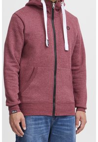 Rode rits hoodie met witte koordjes, zwarte rits en voorzakken. Heeft een textuurstof en een logo detail op de borst.