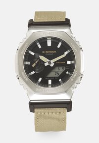 Montre G-Shock avec un boîtier en acier inoxydable argenté, un cadran noir, des accents blancs et dorés, et un bracelet en tissu beige ; résistant à l'eau jusqu'à 20 bars.