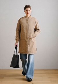 Μπεζ επενδεδυμένο παλτό με ψηλό γιακά και πλαϊνές τσέπες, συνδυασμένο με τζιν παντελόνι φαρδιάς γραμμής και μαύρη τσάντα tote. Φόντο ξύλινο πάτωμα.