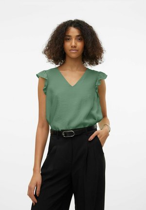 Groene mouwloze blouse met een V-hals en gerimpelde schouderdetails, gecombineerd met hoge zwarte broek en een zwarte riem.