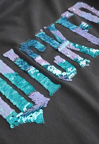 Čierny materiál so žiariacimi teal a fialovými flitrami usporiadanými do tvaru písmen. Flitre sú okrúhle, odrážajú svetlo a vytvárajú textúru.