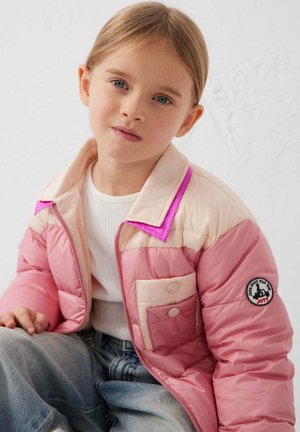 Jong meisje met blauwe ogen, gekleed in een roze en beige gewatteerde jas, een wit geribd shirt en lichtblauwe spijkerbroek, zittend tegen een effen achtergrond.