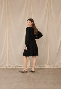 Moteris su ilgais plaukais, vilkinti juodą ilgomis rankovėmis palaidinę ir sijoną, pozuoja šonu priešais smėlio spalvos audinio foną, dėvi zebra rašto aukštakulnius.