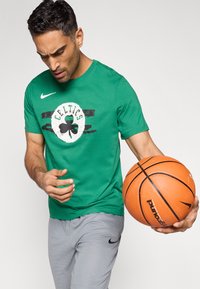 Πράσινο T-shirt με λογότυπο Celtics και μαύρες ρίγες, σε συνδυασμό με γκρι αθλητικό σορτς, κρατώντας ένα πορτοκαλί μπάσκετ. Μάρκα Nike στα ρούχα.