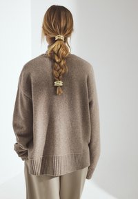 Übergroßer, taupefarbener Pullover mit geripptem Saum und weicher Textur. Lockerer Schnitt, akzentuiert durch einen geflochtenen Pferdeschwanz mit goldenen Haarklammern.