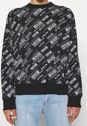 Sort sweatshirt med hvidt diagonalt tekstmønster "Versace Jeans Couture Since 1988", båret med lyseblå jeans.