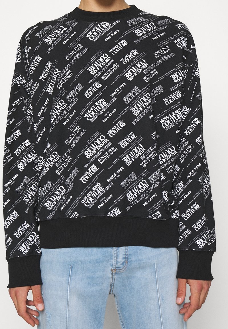 Sweat-shirt noir avec motif texte diagonal blanc « Versace Jeans Couture Since 1988 » porté avec un jean bleu clair.