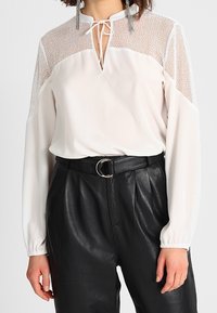 Vit blus med meshpartier vid axlarna, nyckelhålsringning med knytband. Matchas med svarta läderbyxor med hög midja och ett silverbälte.