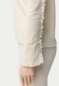 Cremefarbener Pullover mit strukturierten Fransen entlang der Ärmel, gefertigt aus weichem Stoff. Verfügt über eine lockere Passform und einen gerippten Saum.