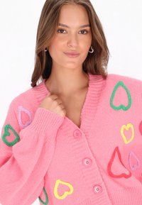 Rosa stickad cardigan med flerfärgade hjärt-mönster, ribbade ärmslut och V-ringning, med två knappar och en mjuk textur.