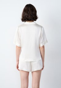 Kobieta z krótkimi brązowymi włosami, ubrana w biały krótki rękaw satynową koszulę oraz dopasowane szorty, stojąca przodem do gładkiej białej ściany.