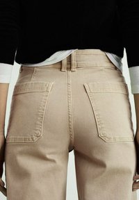 Jeans in denim beige con vita alta, tasche posteriori e una consistenza liscia, abbinati a un maglione nero con dettagli bianchi sulle maniche.