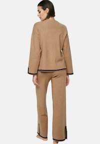 Tweed set in camelkleurige stof. Gebreide trui met lange mouwen en zwarte afwerking, gecombineerd met bijpassende wijde broek met zijsplitten.